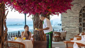 A seaside restaurant with outdoor seating, a waiter serving a meal, and a view of the ocean. - Playa Los Arcos Hotel Beach Resort & Spa (Puerto Vallarta)