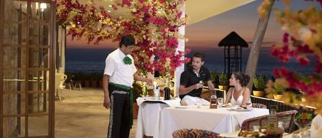 A restaurant with a view of the sea, tables set with white tablecloths, and a waiter serving guests.
