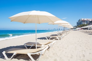 A beach with rows of white sun loungers and umbrellas, a clear blue sky, and a cityscape in the background.