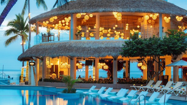 A resort with a pool, lounge chairs, and a thatched-roof building with hanging lights. - Playa Los Arcos Hotel Beach Resort & Spa (Puerto Vallarta)