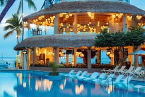 A resort with a pool, lounge chairs, and a thatched-roof building with hanging lights.