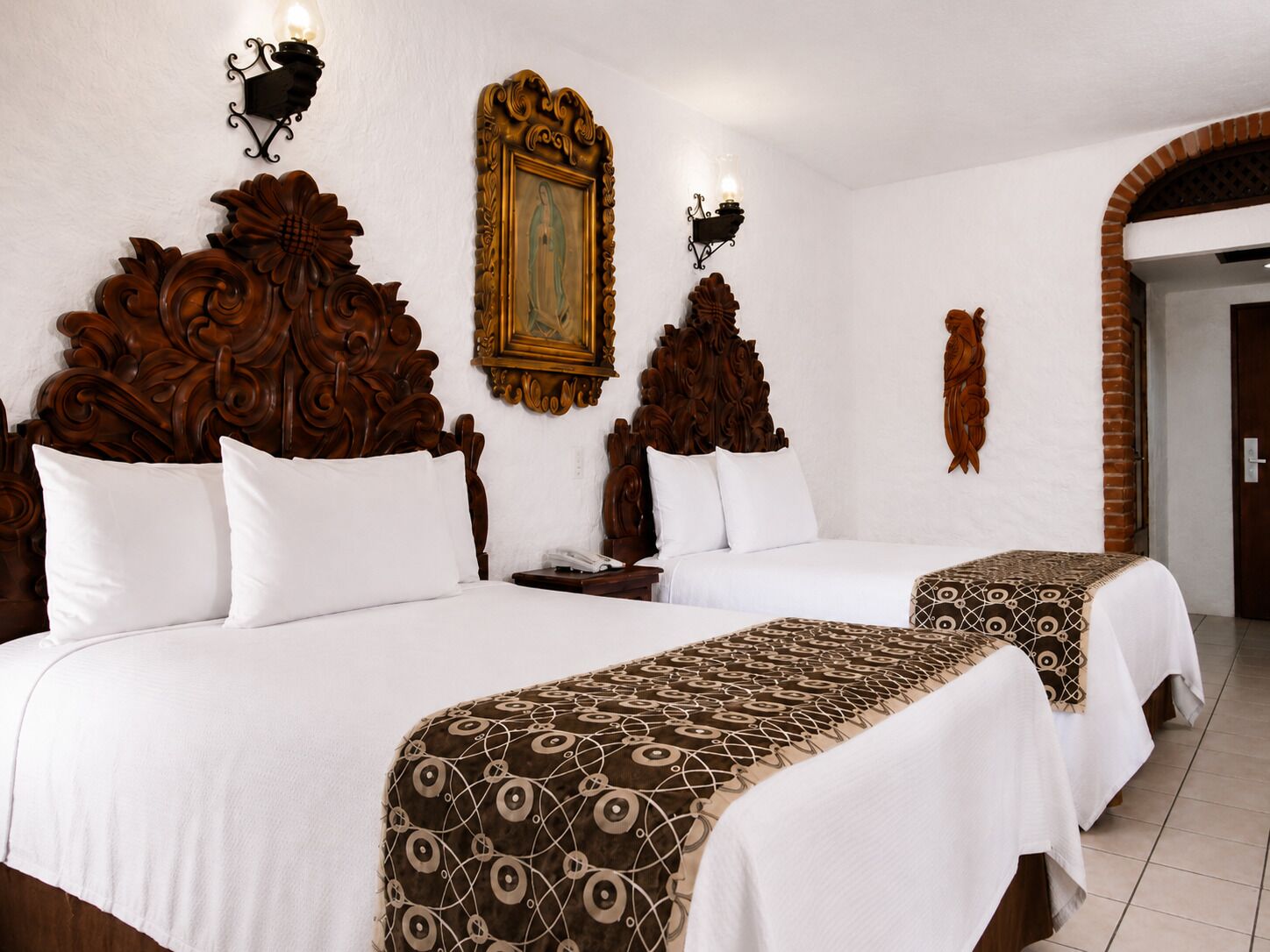 A hotel room with two beds, ornate wooden headboards, a framed painting, wall sconces, and a brick archway.