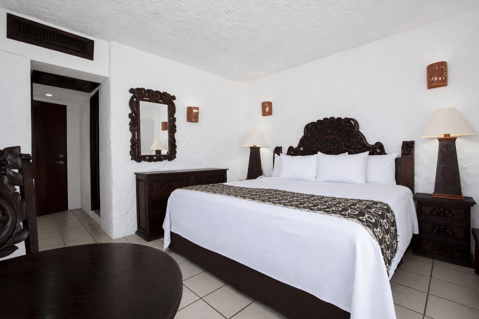 A hotel room with a large bed, a wooden headboard, a dresser, a mirror, two bedside lamps, and a small table.
