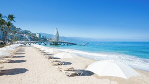 A beach with rows of sun loungers and umbrellas, a pier extending into the ocean, and buildings in the background. - Playa Los Arcos Hotel Beach Resort & Spa (Puerto Vallarta)