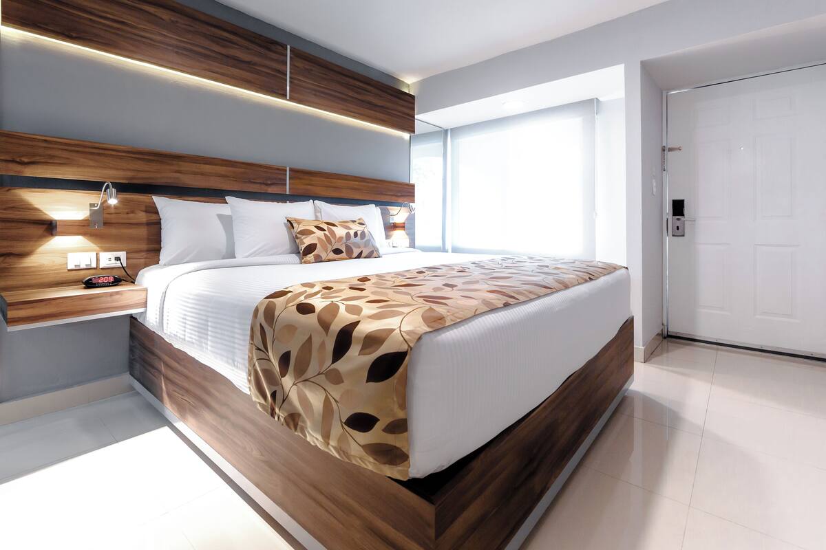 A modern bedroom with a wooden headboard, a large bed with patterned bedding, a bedside table with a lamp, and a large window providing natural light.