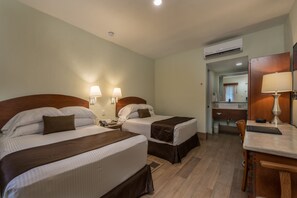 Habitación de hotel con dos camas, un escritorio, una silla, una lámpara y un baño visible a través de una puerta abierta.