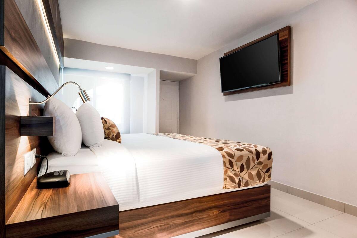 A modern hotel room with a wooden bed frame, a flat-screen TV mounted on the wall, and a bedside table with a lamp and a remote control.