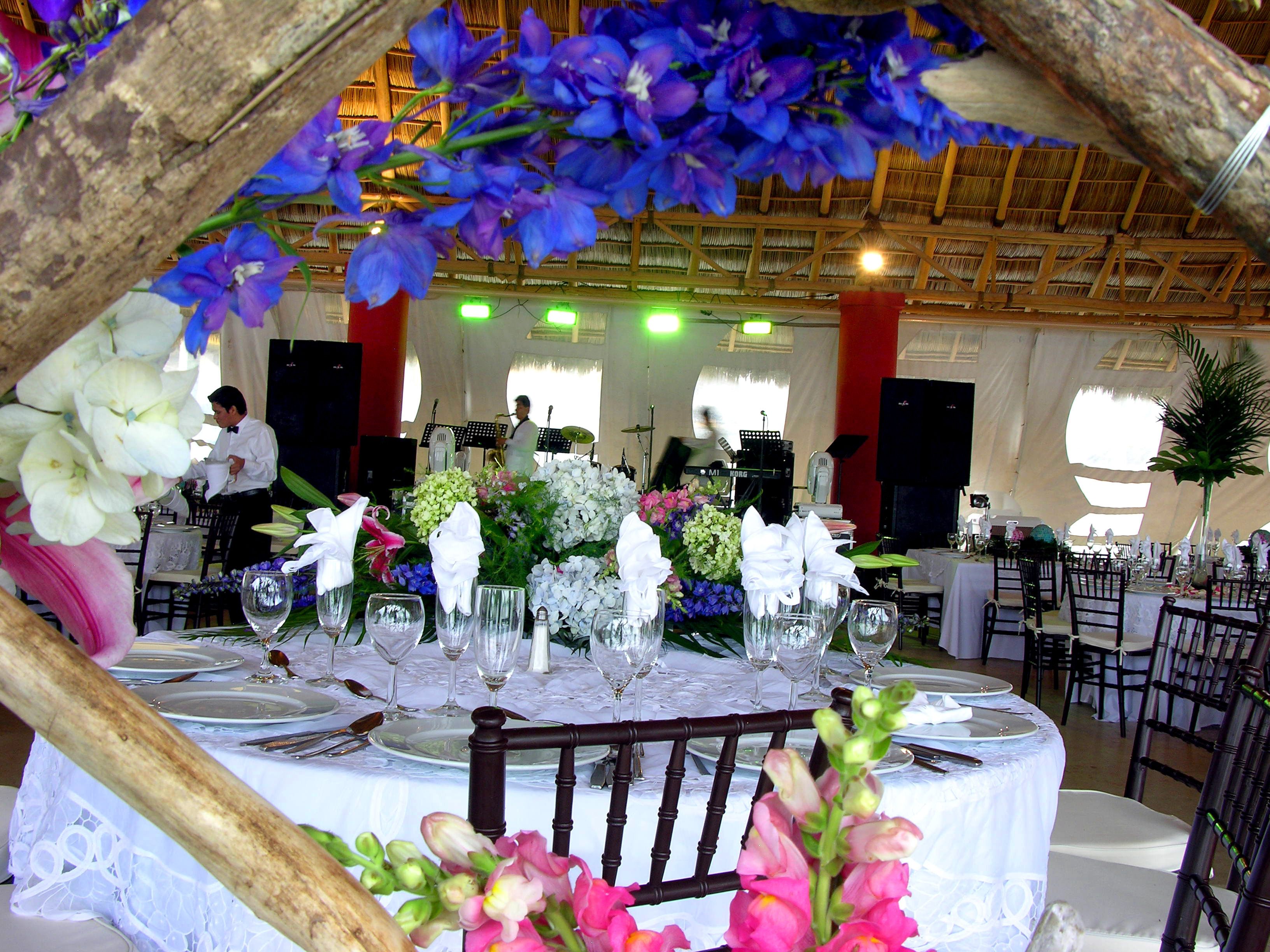 Un salón de banquetes con mesas dispuestas para un evento formal, destacando centros de mesa florales y una banda en el fondo.