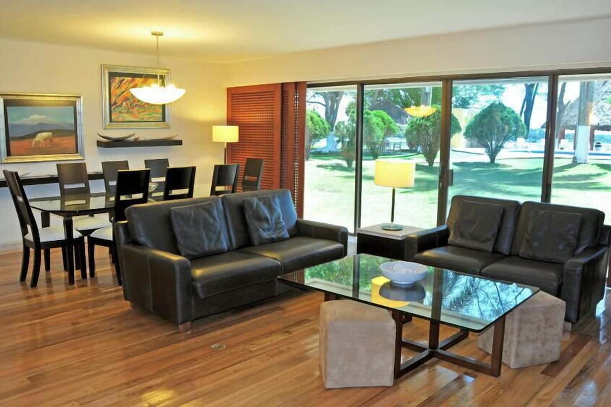 A modern living room with a glass coffee table, two sofas, a dining table with chairs, and large windows offering a view of a garden.