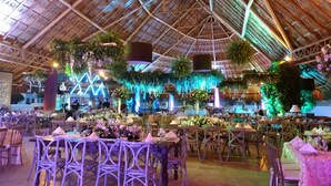 Un espacio interior con techo de paja, mesas dispuestas con centros de flores y plantas colgantes.