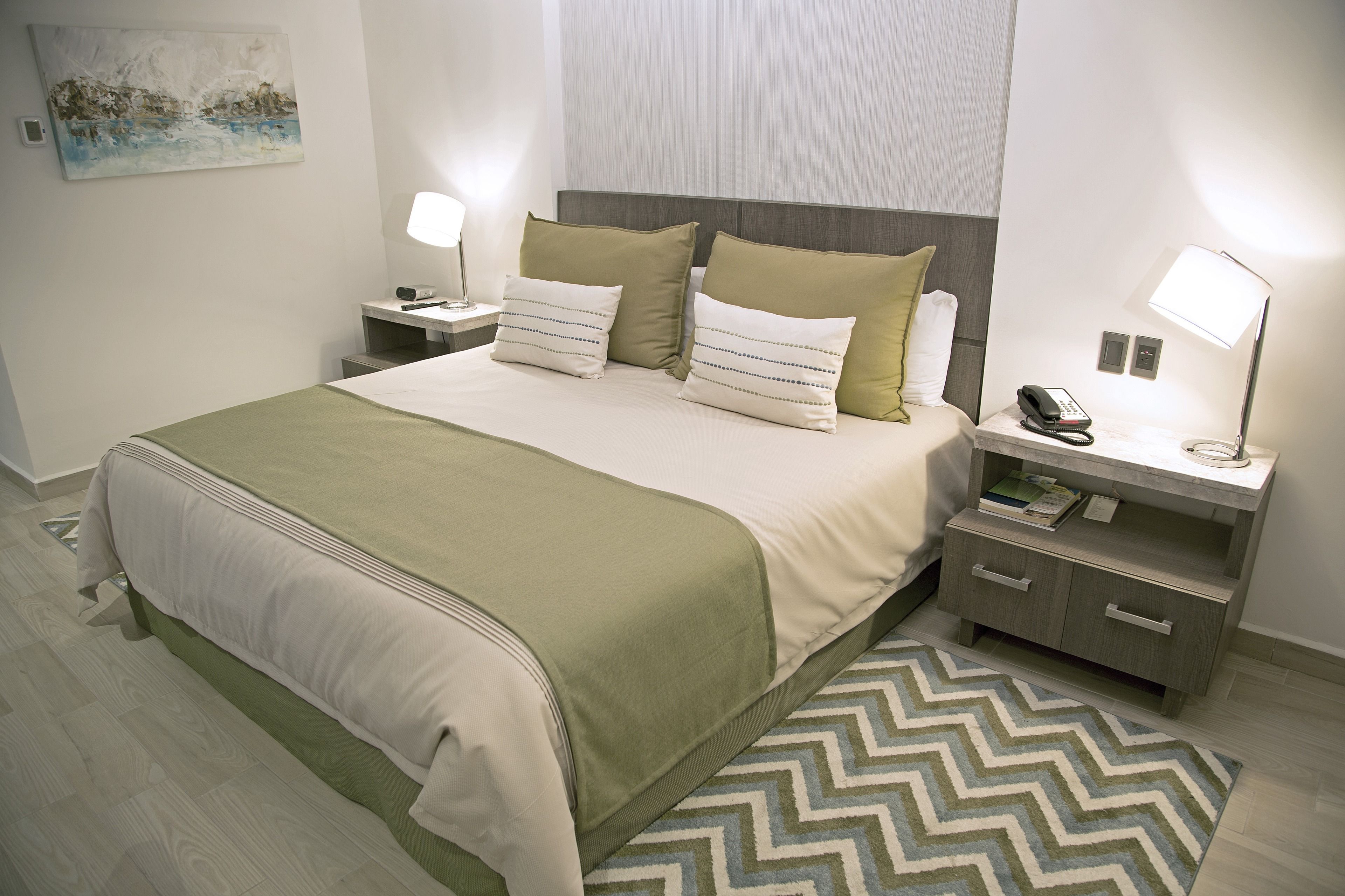 A neatly arranged hotel room with a bed, two bedside tables, a nightstand, a lamp, a phone, and a painting on the wall.