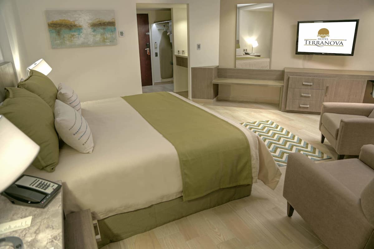 A hotel room with a bed, a desk with a lamp, a TV, a mirror, and a bathroom visible through an open door.