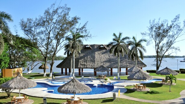 Un resort con zona de piscina, construcciones de techo de paja, palmeras y vista al agua.