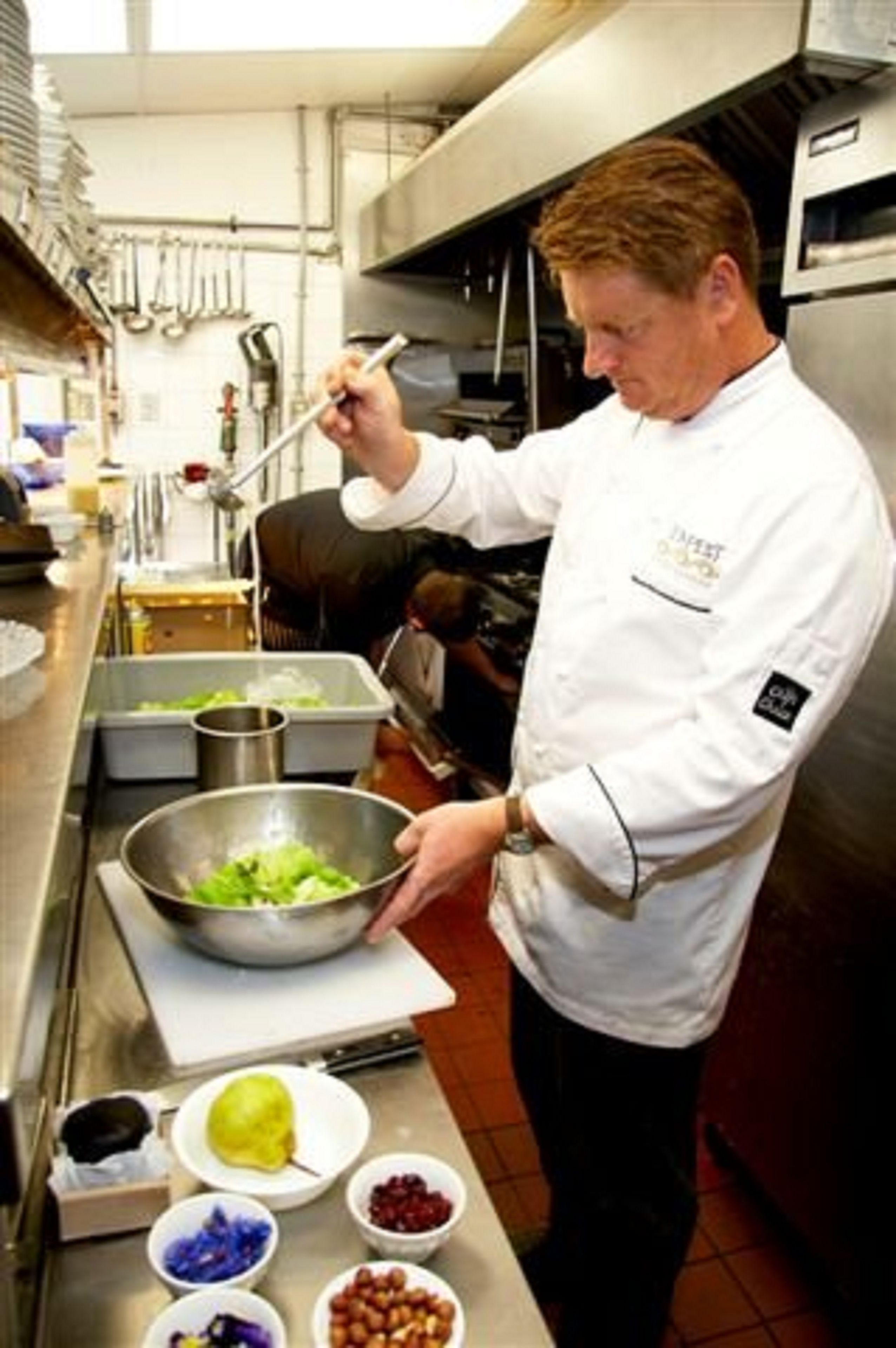 Een kok bereidt eten in een professionele keuken met roestvrijstalen oppervlakken, diverse kookgerei en ingrediënten zoals groenten en fruit.