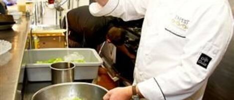 Een kok bereidt eten in een professionele keuken met roestvrijstalen oppervlakken, diverse kookgerei en ingrediënten zoals groenten en fruit.