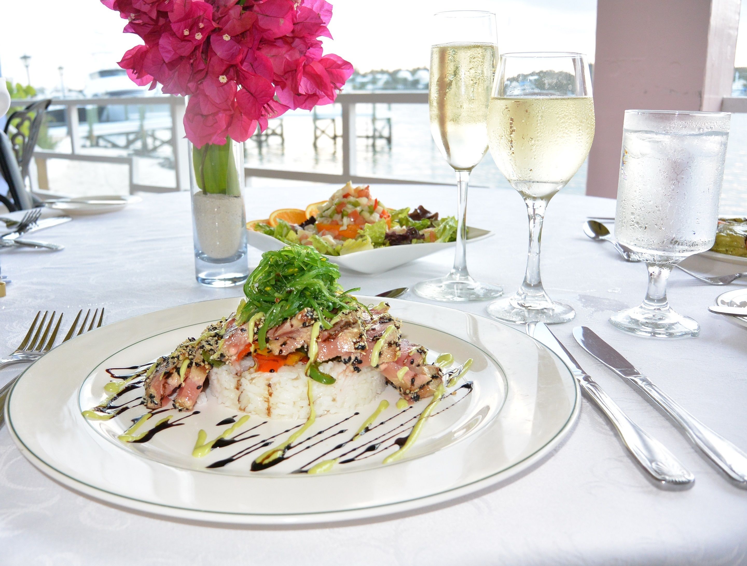 A dining table with a plate of food, two glasses of white wine, and a vase of pink flowers.