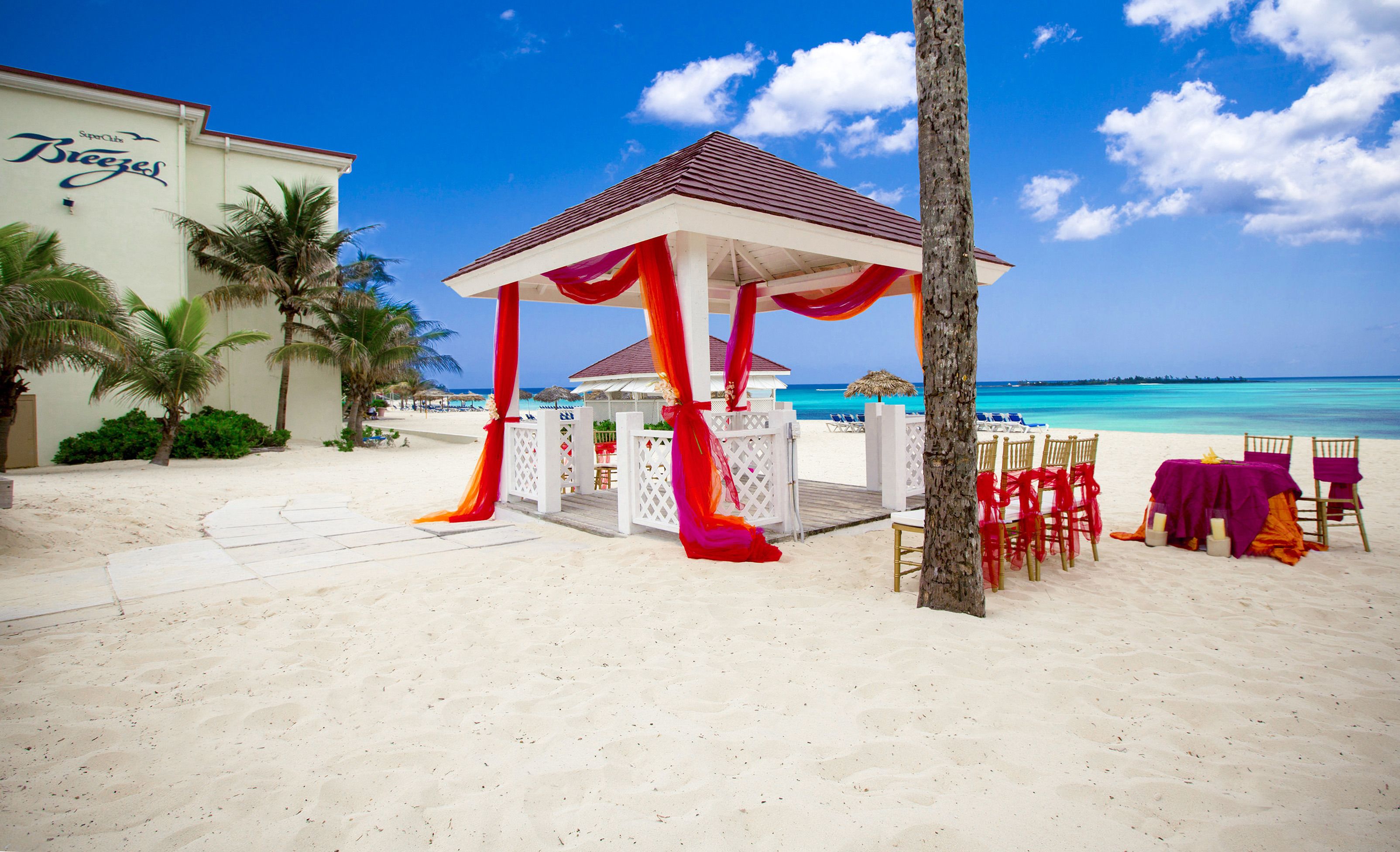 A beach gazebo with red drapes, a white building with 'Breezes' signage, and a clear blue sky.