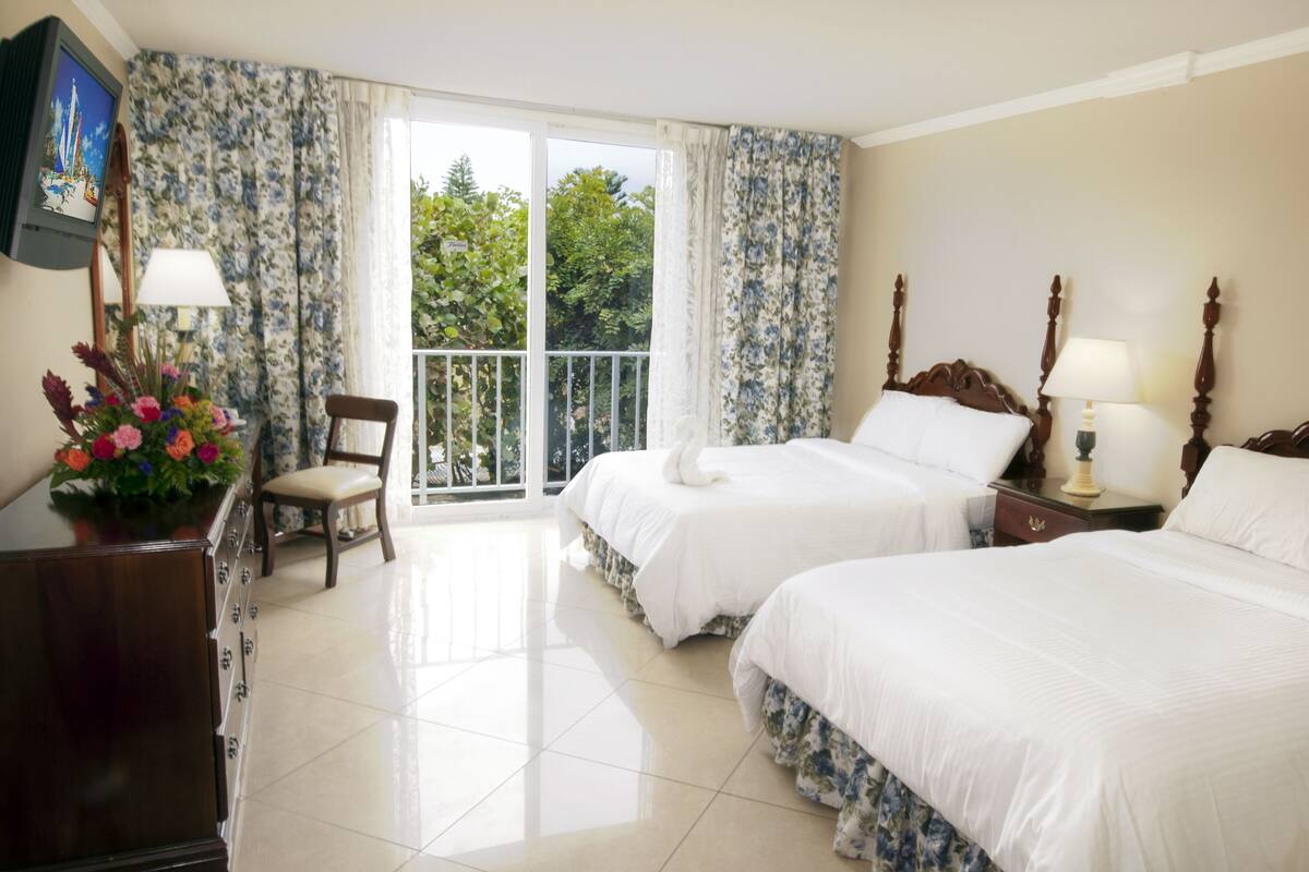 A hotel room with two beds, a TV, a chair, a desk, and a balcony with a view of greenery.