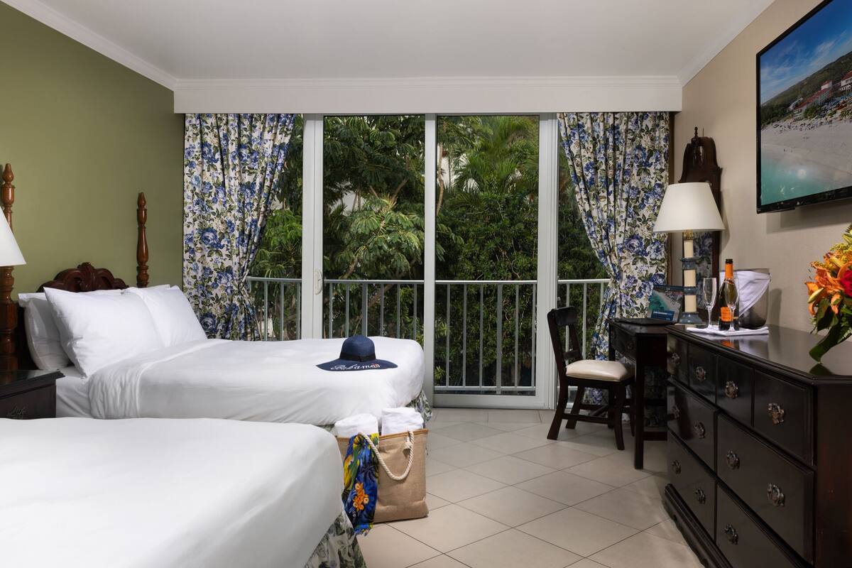 A hotel room with two beds, a desk with a lamp, a chair, a dresser, and a large window with curtains.