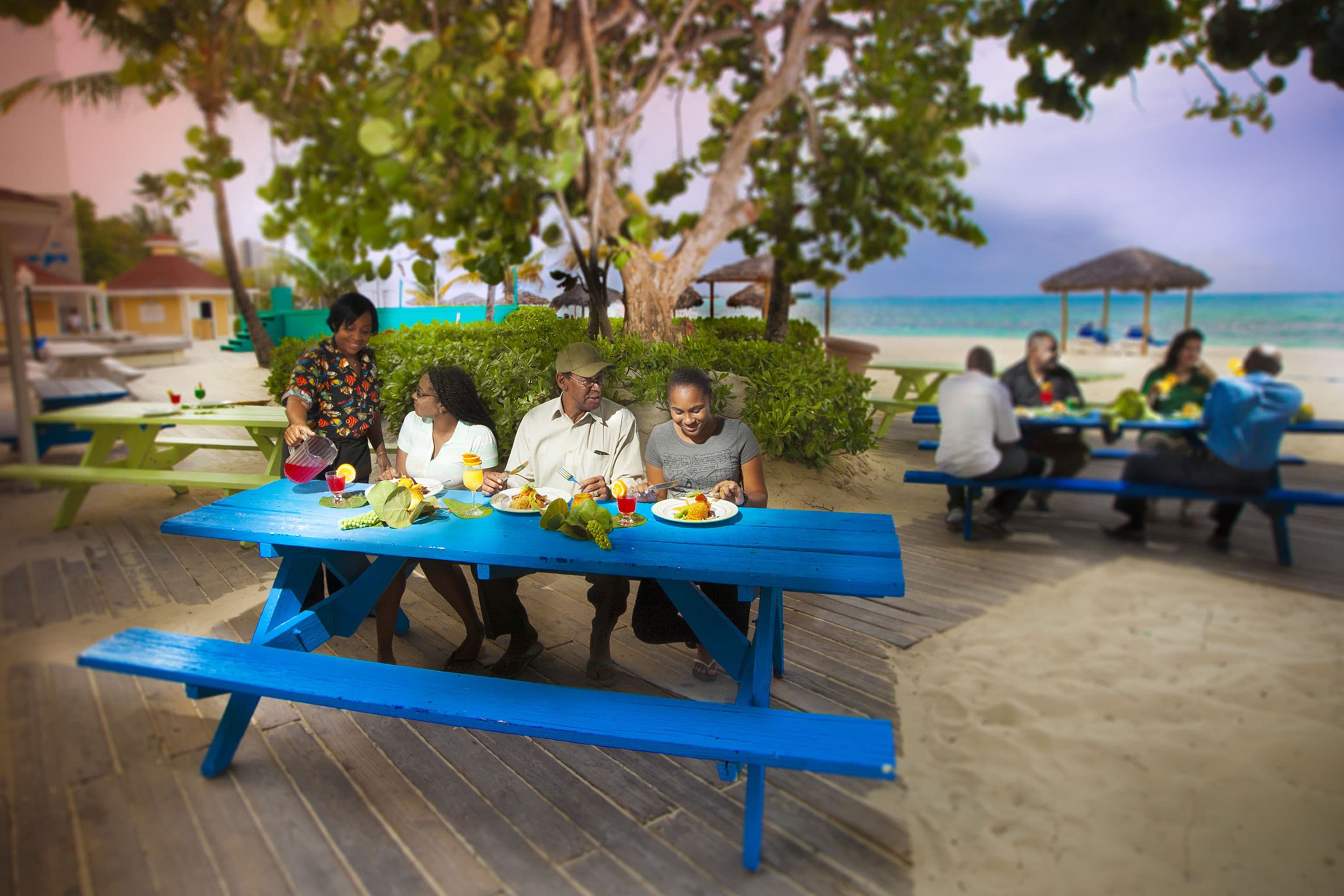A group of people dining at a blue picnic table on a wooden deck with a beach and ocean view in the background.