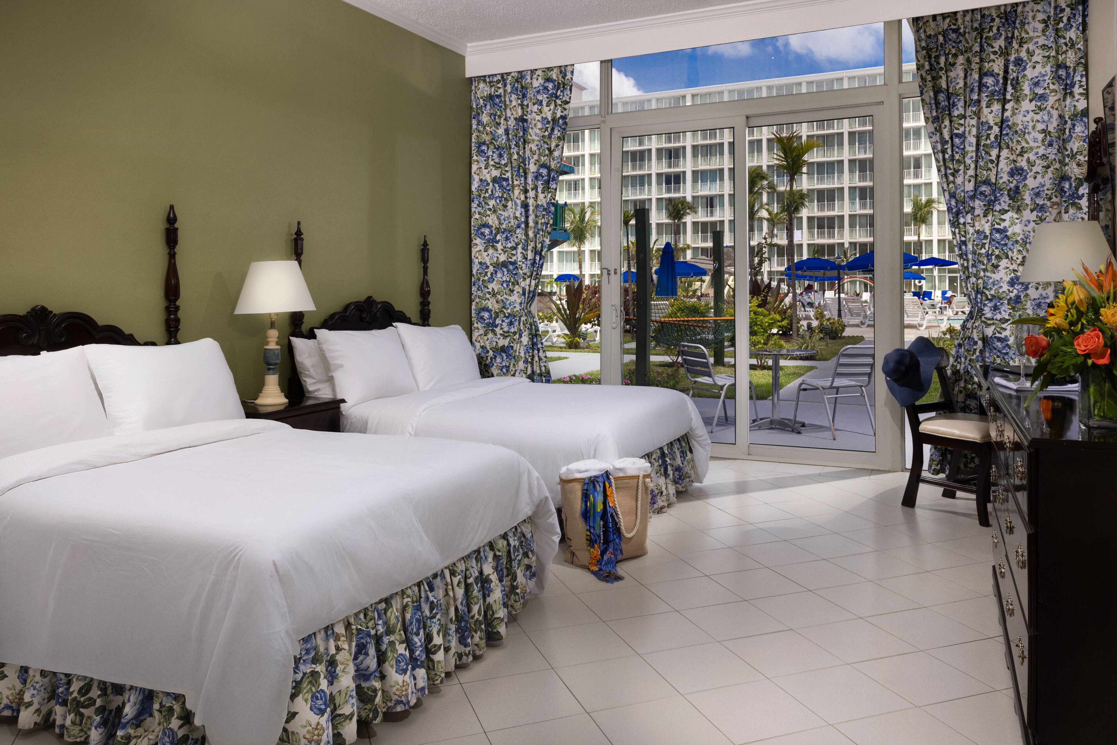 A hotel room with two beds, a balcony with a view of a pool and building, and a desk with a chair.