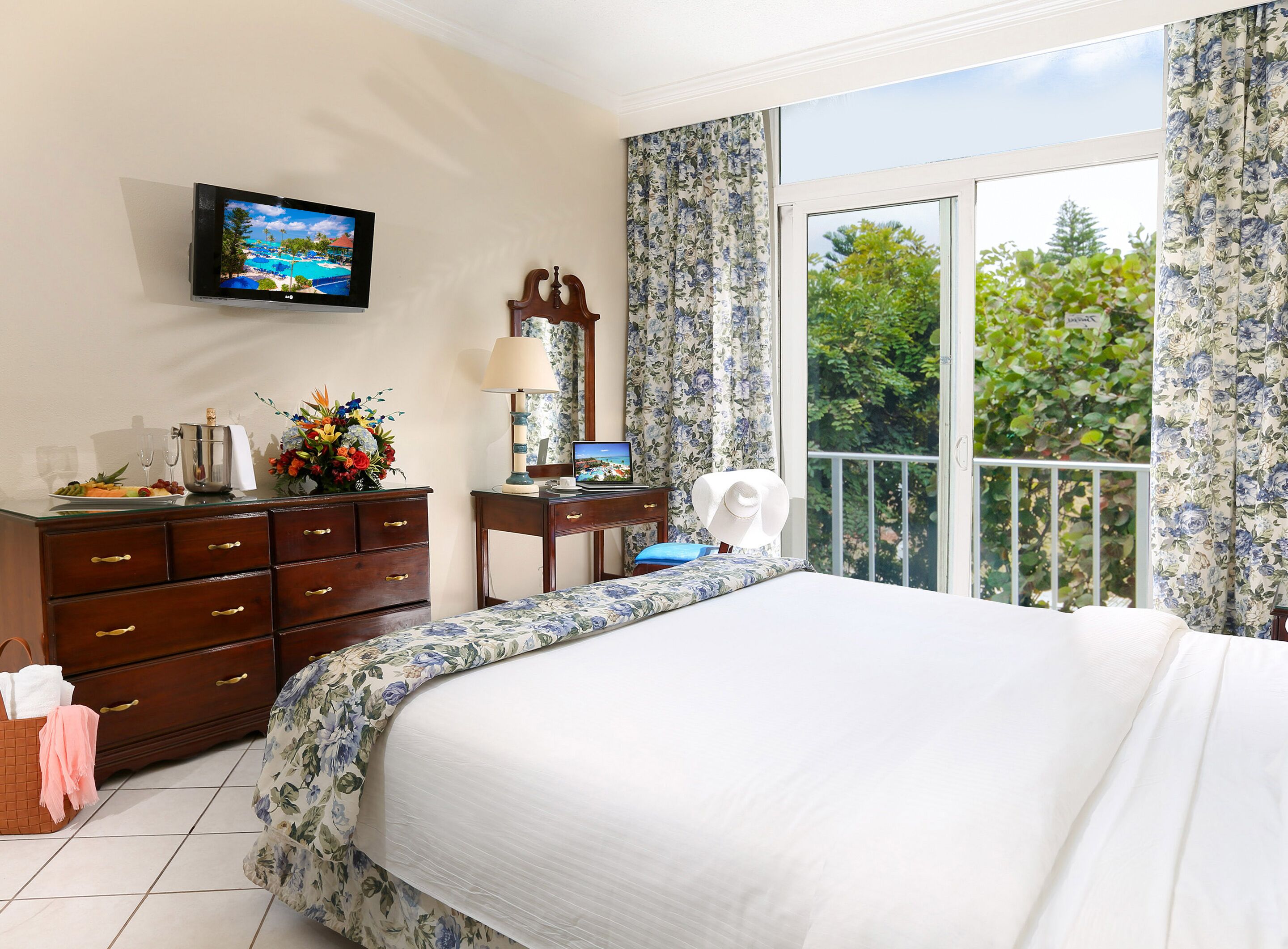 A hotel room with a bed, a desk with a laptop, a TV, and a balcony with a view of trees.
