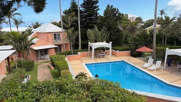 A swimming pool with lounge chairs, a covered patio, and a building with a sloped roof.