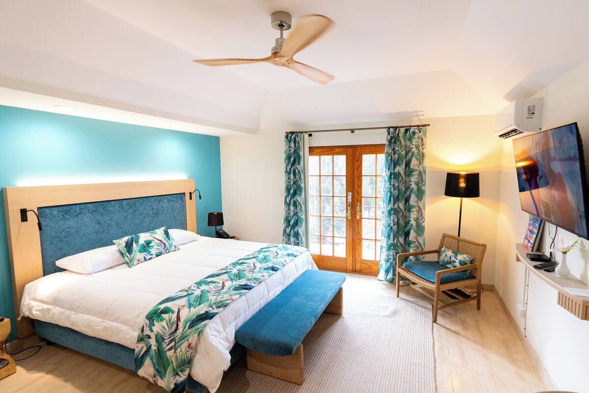 A bedroom with a bed, a bench, a chair, a ceiling fan, a TV, and a window with curtains.