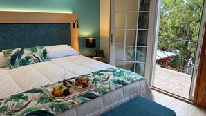 A hotel room with a bed, a bedside table with a lamp, a tray of fruit, and a view of trees through a glass door. - Fourways Inn (Southampton Parish)