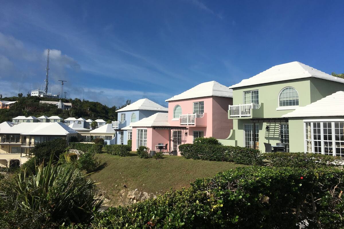 A row of colorful houses with white roofs, surrounded by greenery and a clear blue sky.