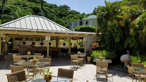 An outdoor patio area with wicker furniture and glass tables, surrounded by lush greenery and a white building in the background. - Blue Horizons Garden Resort (St. George's)