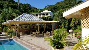 A pool area with a covered seating area, lounge chairs, and a building with a white roof in the background. - Blue Horizons Garden Resort (St. George's)