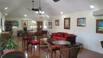 A spacious living area with a red sofa, multiple chairs, a coffee table, and a ceiling fan. There are bookshelves, a potted plant, and artwork on the walls.