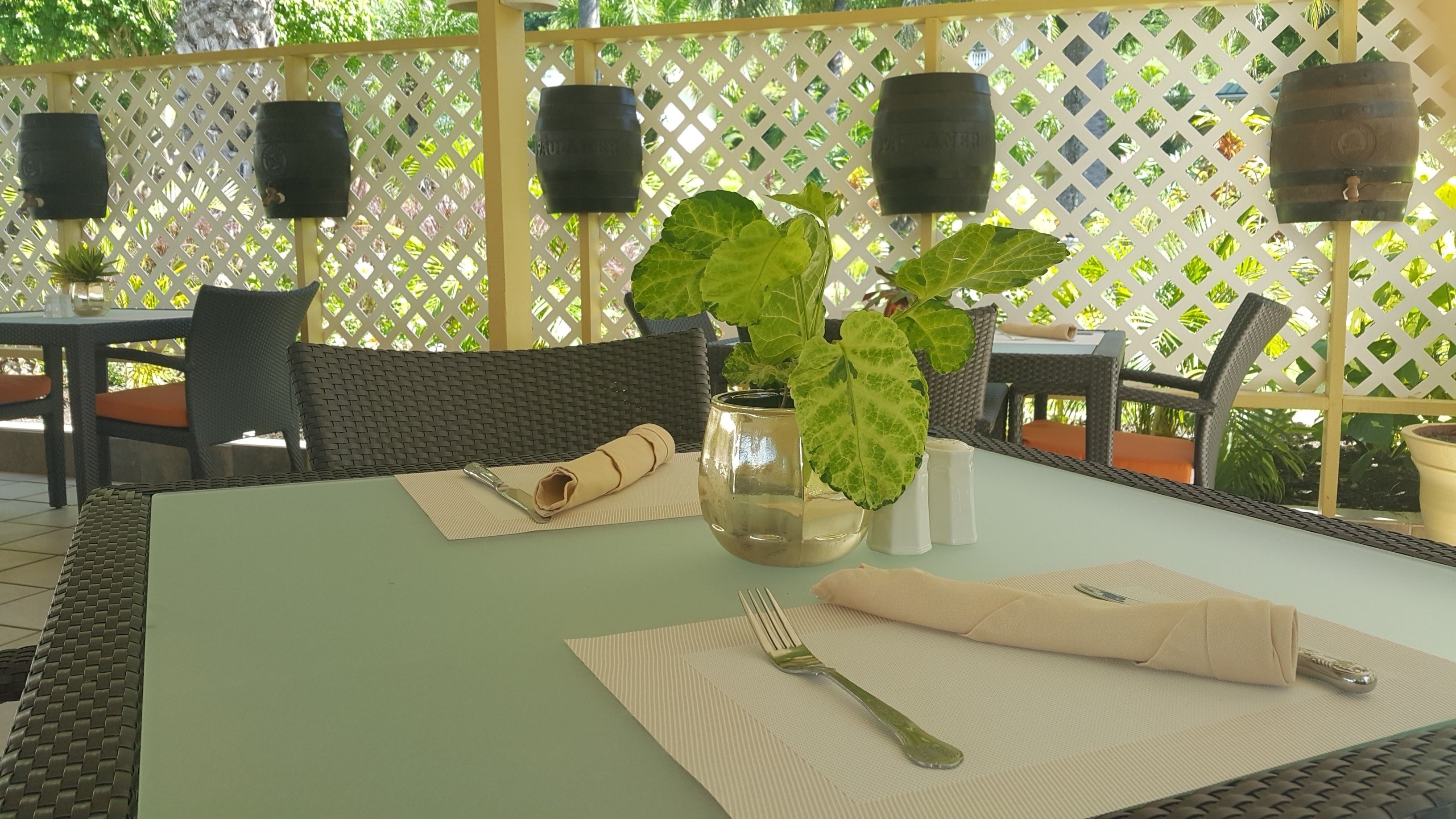 An outdoor dining area with wicker chairs, a table set with a folded napkin, a fork, a salt shaker, and a potted plant.