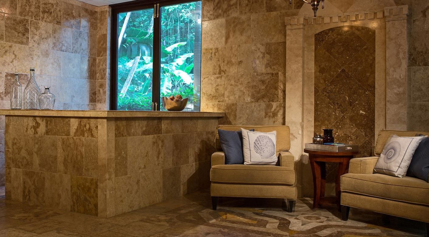 A room with a stone wall, a window with a view of greenery, two armchairs, and a small table.