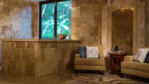 A room with a stone wall, a window with a view of greenery, two armchairs, and a small table.