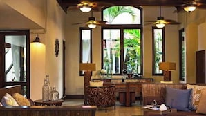 A spacious living area with a sofa, coffee table, and a dining table with chairs. There are large windows with a view of greenery outside. - Carambola Beach Resort (Kingshill)