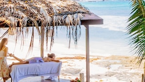 A beachside massage area with a thatched roof, a massage table, and a person receiving a massage.