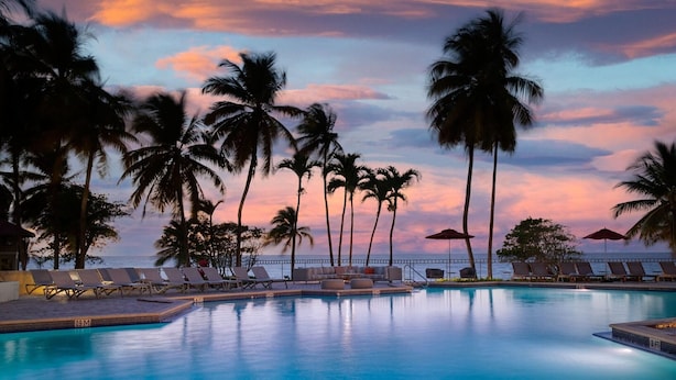 A poolside area with lounge chairs and palm trees, overlooking a body of water. - Carambola Beach Resort (Kingshill)
