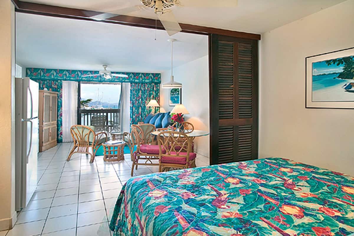 A hotel room with a bed, a balcony with wicker chairs, a dining table, and a ceiling fan.