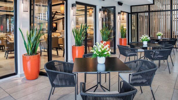 A modern indoor dining area with glass doors leading to a kitchen, black wicker chairs, and potted plants. - Real InterContinental San Salvador at Metrocentro Mall by IHG (San Salvador)