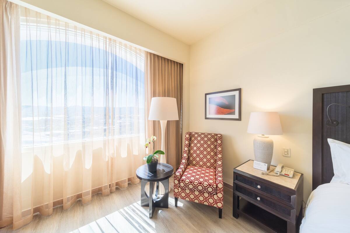Une chambre d’hôtel avec une grande fenêtre, une chaise, une petite table avec une lampe, une table de chevet avec une lampe et un lit.