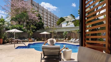 Zona junto a la piscina con sillas de descanso, una mesita con una bebida y vista a un edificio al fondo.