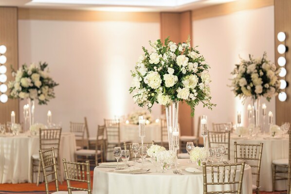 Un salón de banquetes con mesas redondas dispuestas para un evento formal, con centros de mesa florales blancos y candelabros.