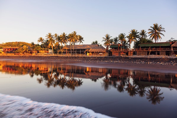 A beachfront hotel with palm trees, a thatched-roof structure, and a calm reflective water surface.