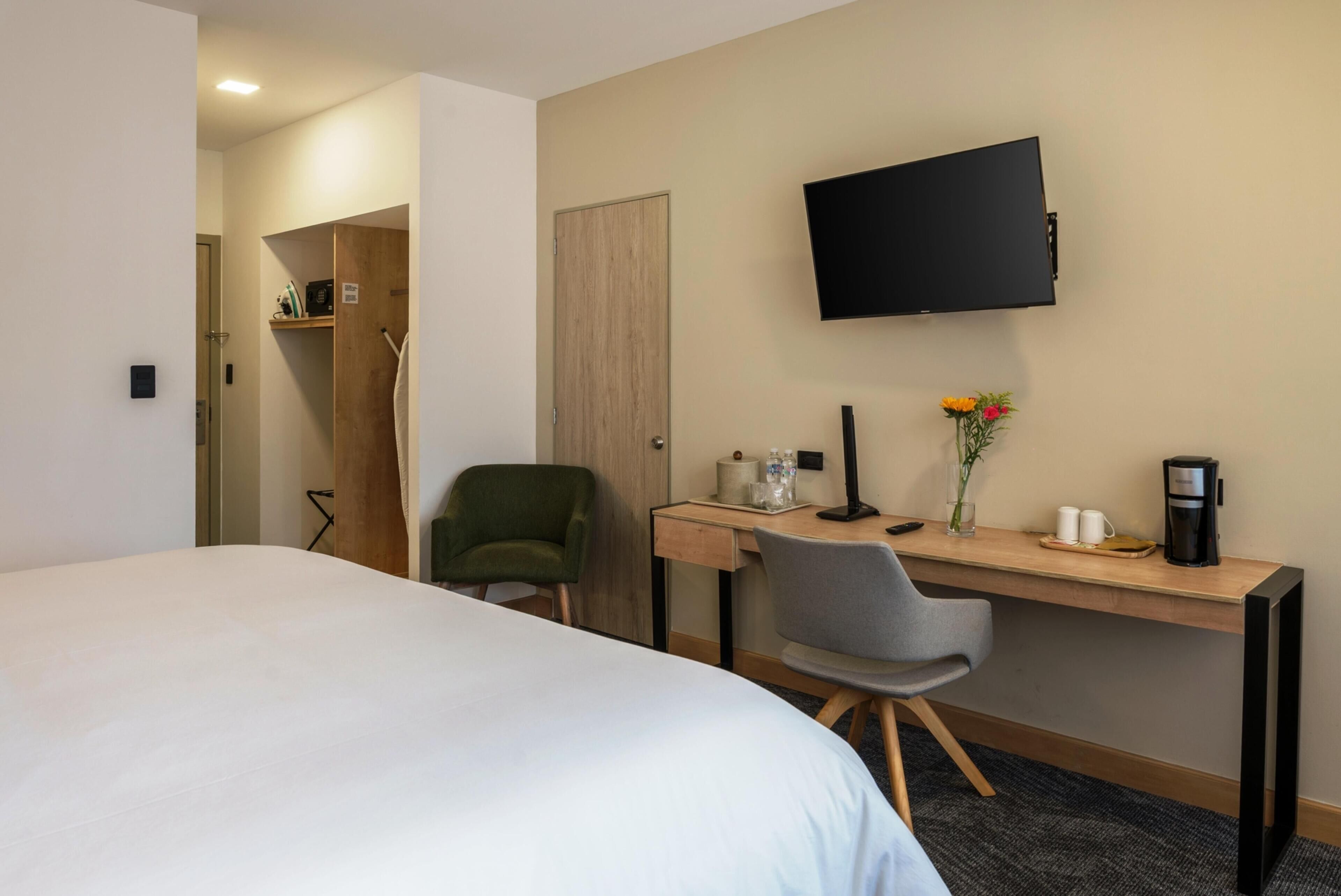 A hotel room with a bed, a desk with a chair, a TV, a coffee maker, and a vase with flowers.