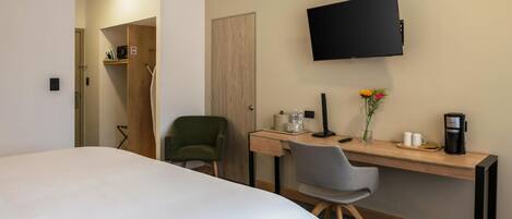 A hotel room with a bed, a desk with a chair, a TV, a coffee maker, and a vase with flowers.