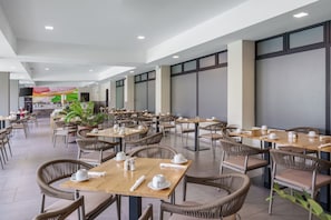 Un vaste espace intĂ©rieur amĂ©nagĂ© en salle Ă manger, avec de nombreuses tables et chaises, une cuisine Ă lâarriĂšre-plan et de grandes fenĂȘtres laissant entrer la lumiĂšre naturelle.