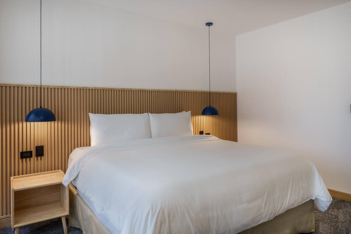 Cama bem arrumada com cabeceira de madeira, duas luminárias de cabeceira e uma pequena mesa de cabeceira de madeira.