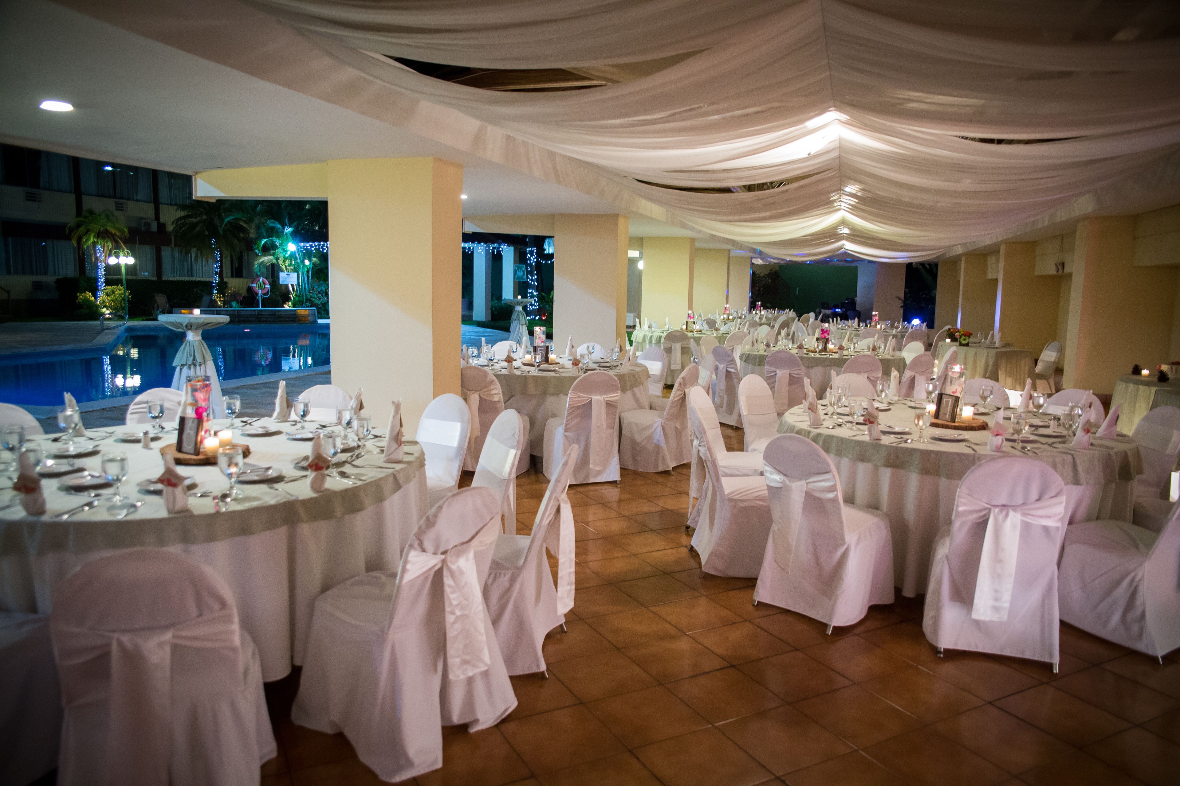 Un salón de banquetes con mesas redondas dispuestas para un evento formal, con manteles blancos y sillas con cintas rosas.